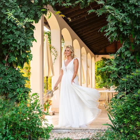 Die Braut steht im weißen Kleid und mit einem Blumenstrauß in der Hand am Rande einer Terrasse, mit Arkaden umgeben.